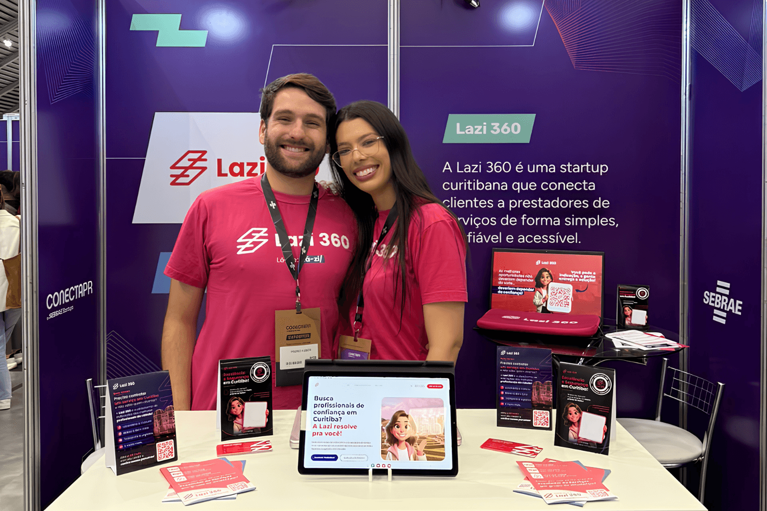 Larissa Rocha e Pedro Huber em seu estande no ConectaPR 2026 atrás de uma mesa com um tablet com o site da Lazi 360 aberto e flyers.